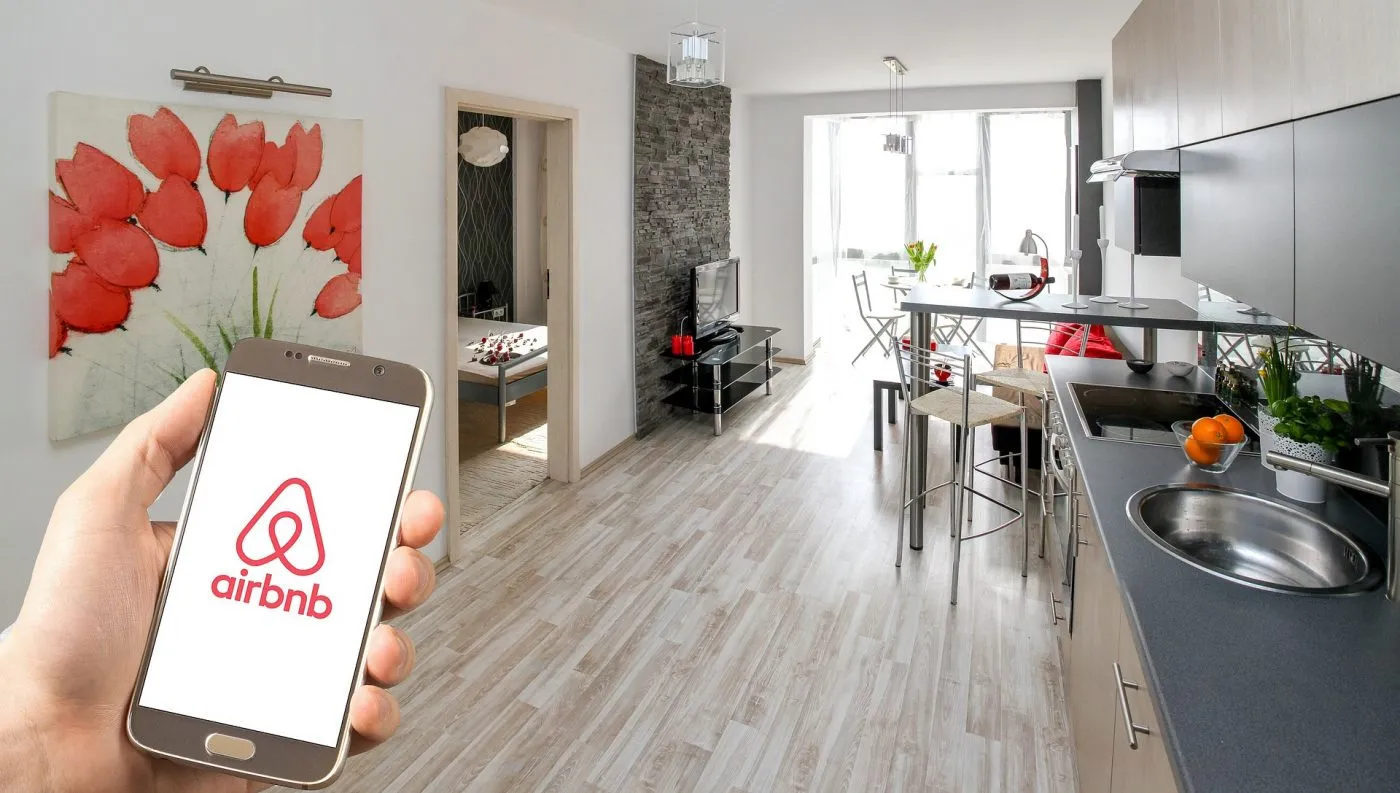 Una mano sostiene un teléfono móvil que muestra en su pantalla el logotipo de Airbnb, con el interior de un apartamento moderno y luminoso de fondo.