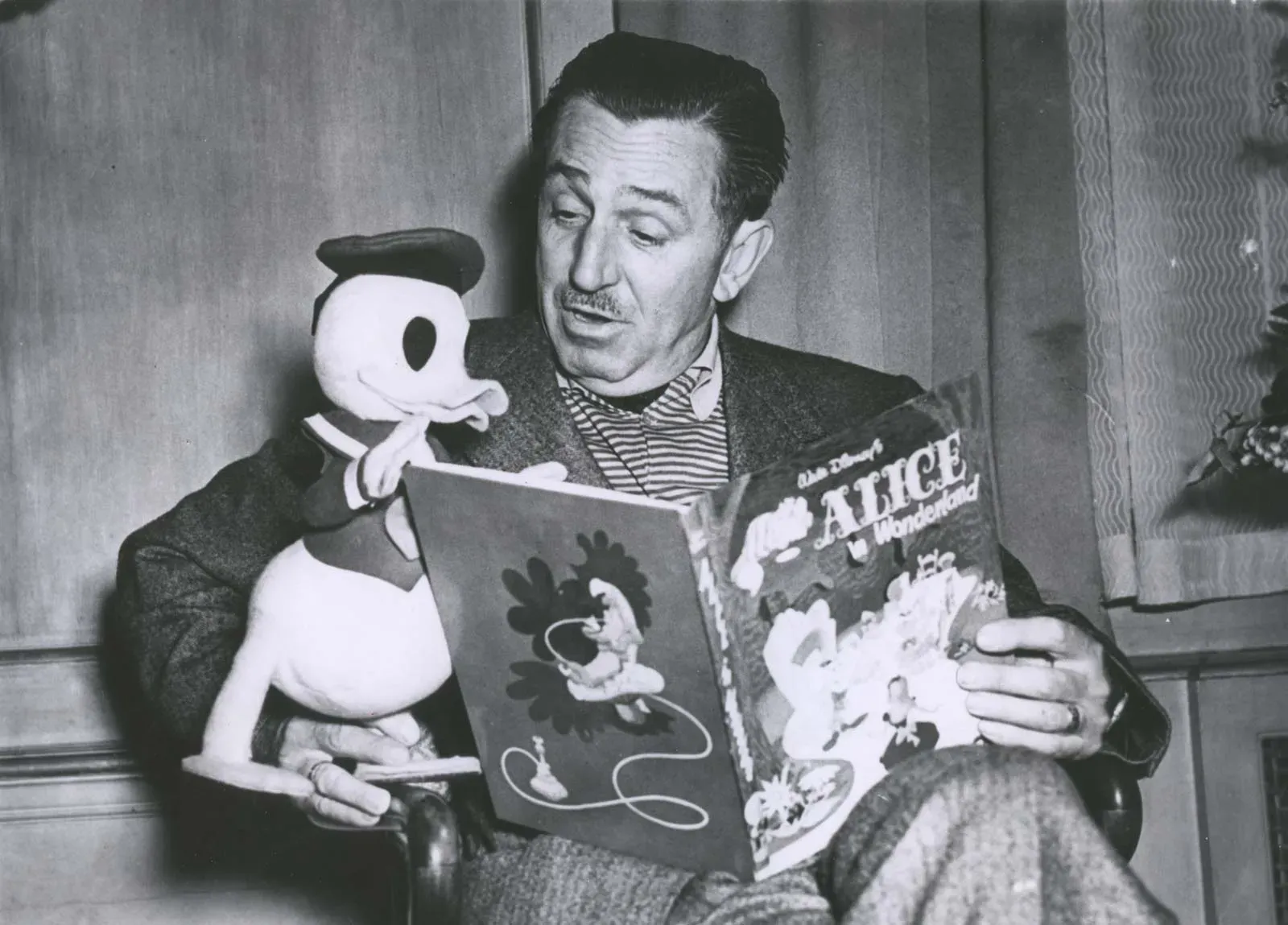 Fotografía antigua en blanco y negro de Walt Disney sentado, sosteniendo un muñeco del Pato Donald mientras leen juntos un libro ilustrado de "Alicia en el país de las maravillas".